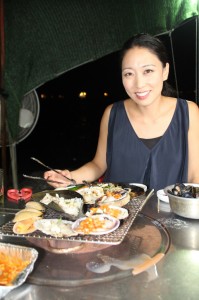 Chef Judy Joo sits down for some grilled clams by the sea near Taejondae Resort Park in Busan for her new series "Korean Food Made Simple." Photo courtesy of  Blink Films/ Media Story 9 Prods